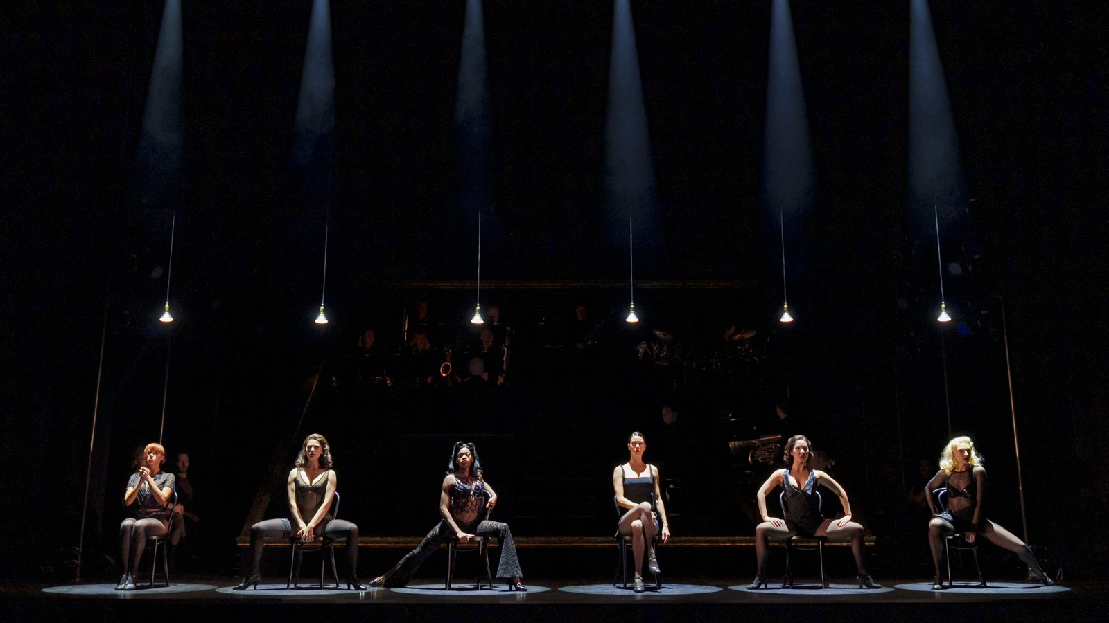 Six Chicago performers seated across a dark stage beneath individual hanging lights.