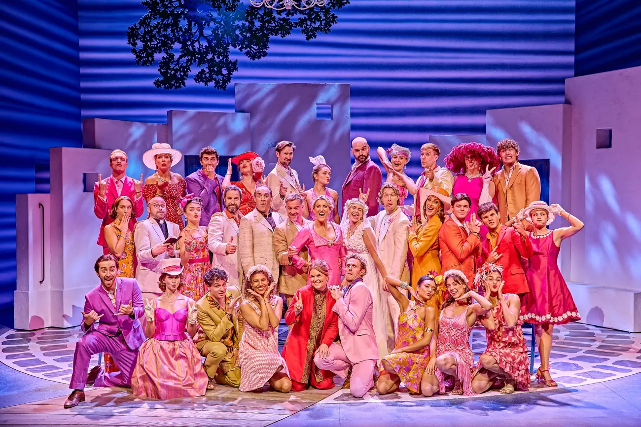 The Mamma Mia! company posing together on stage in colourful costumes.