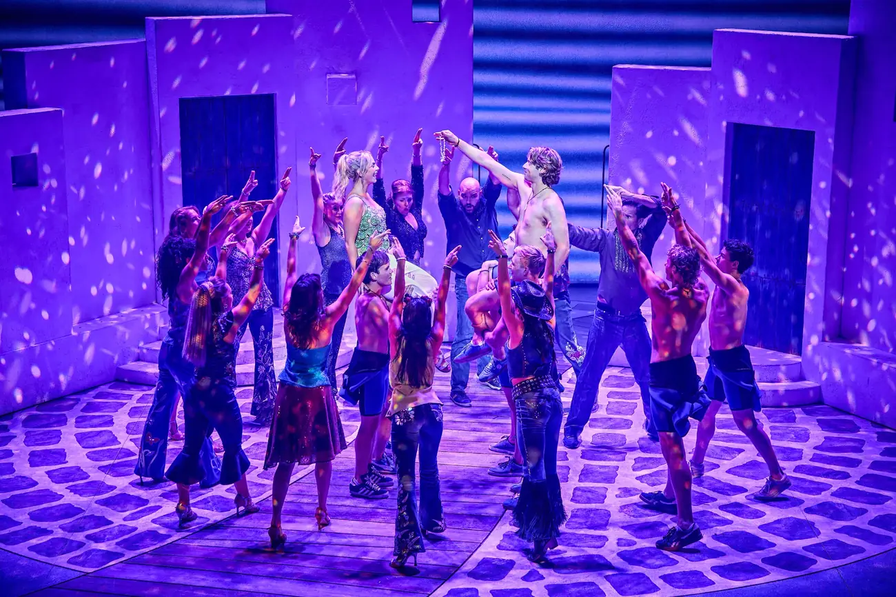 The Mamma Mia! company performing a dance sequence under purple and blue stage lighting.
