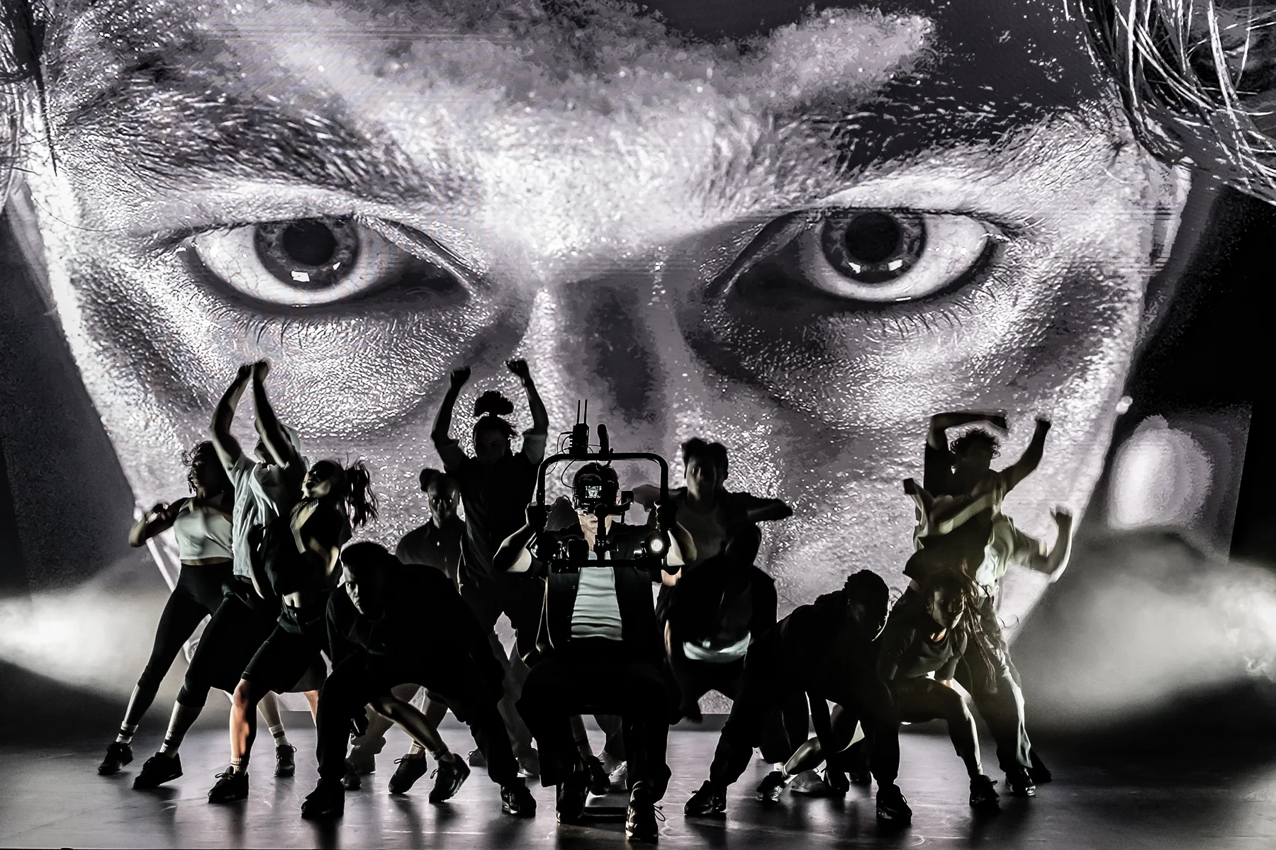 The Sunset Boulevard company performing in front of a large black-and-white projected face.